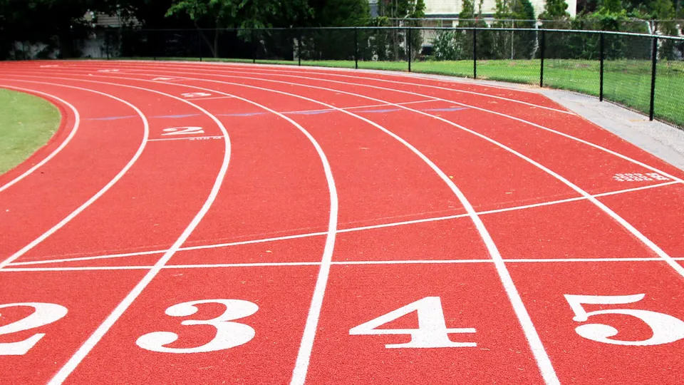 Red running track with white lane markings and numbers, curving past a fence and trees, daytime